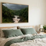 A bed with oatmeal and green linen bedding under one large framed rainforest print in a thin natural wood frame, showing layered fronds and mist between trees in muted greens and soft b