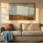 A quiet living room at golden hour: an oat linen sofa against a warm off-white wall, a dusty sky blue throw draped over one arm, a single clay-coloured cushion, and a terracotta pot