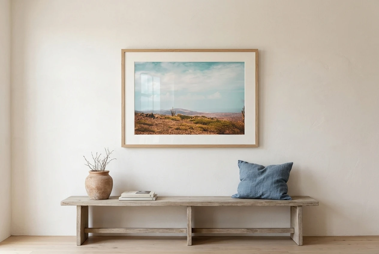 A single framed print of clay-coloured desert hills under a pale blue sky, in a natural oak frame with a wide warm white mat, hung with generous bare wall around it above a console where a terracotta pot sits beside a blue linen cushion.  please include the referenced image as the wall are.