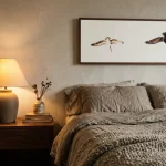 A bedside arrangement in stone and oatmeal tones: rumpled linen sheet edge, chunky wool throw folded at the bed's foot, a walnut nightstand with a matte ceramic lamp base and a small